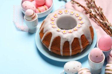 Delicious Easter cake decorated with sprinkles near painted eggs and willow branches on light blue background