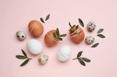 Beautifully decorated Easter eggs and green leaves on pale pink background, flat lay