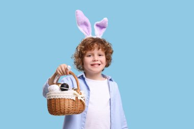Portrait of happy boy in cute bunny ears headband holding wicker basket with Easter eggs on light blue background