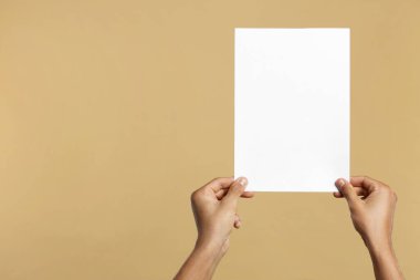 Man holding sheet of paper on beige background, closeup. Mockup for design