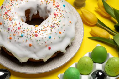 Easter cake with sprinkles, painted eggs and tulips on yellow background