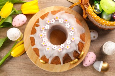 Delicious Easter cake decorated with sprinkles near painted eggs and tulips on wooden table, flat lay