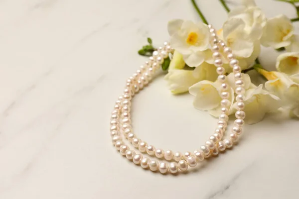 Beautiful pearl necklace and flowers on white marble table, closeup. Space for text