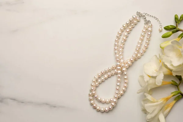 Beautiful pearl necklace and flowers on white marble table, flat lay. Space for text