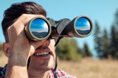 Man looking through binoculars outdoors on sunny day. Mountain landscape reflecting in lenses