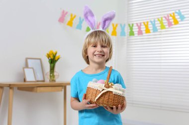 Happy boy in bunny ears headband holding wicker basket with painted Easter eggs indoors