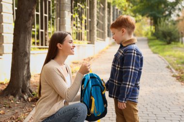 Young mom giving school backpack to her son outdoors