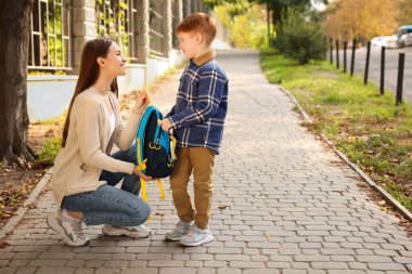 Young mom giving school backpack to her son outdoors