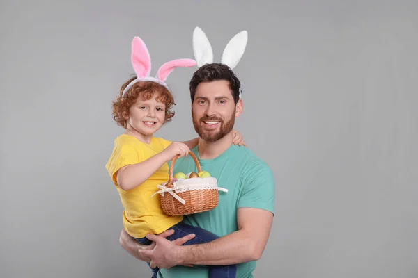 Happy father and son wearing cute bunny ears headbands on light grey background. Boy holding Easter basket with painted eggs