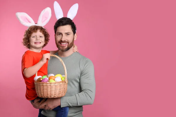 Happy father and son wearing cute bunny ears headbands on pink background. Boy holding Easter basket with painted eggs, space for text