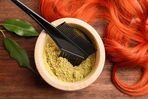 Bowl of henna powder, brush, green leaves and red strand on wooden table, flat lay. Natural hair coloring