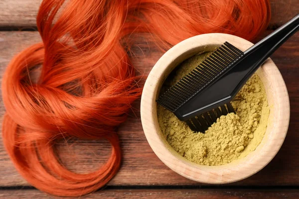 Bowl of henna powder, brush and red strand on wooden table, flat lay. Natural hair coloring