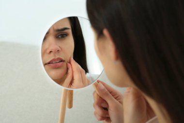 Young woman looking in mirror on her pimples indoors. Hormonal disorders