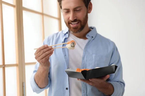 Happy man eating sushi rolls with chopsticks indoors, focus on hand