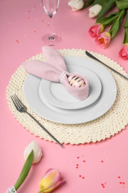 Festive table setting with painted egg, plates and tulips on pink background. Easter celebration