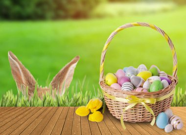 Bunny hiding in green grass near wooden deck with wicker basket full of Easter eggs