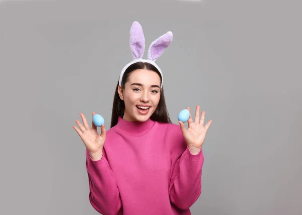 Happy woman in bunny ears headband holding painted Easter eggs on grey background