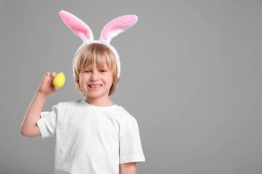 Happy boy in bunny ears headband holding painted Easter egg on grey background. Space for text