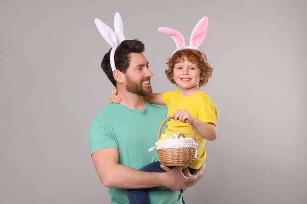 Happy father and son wearing cute bunny ears headbands on light grey background. Boy holding Easter basket with painted eggs