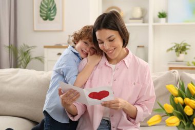 Little son congratulating his mom with Mother`s day at home. Woman holding handmade greeting card