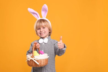 Happy boy in bunny ears headband holding wicker basket with painted Easter eggs on orange background. Space for text