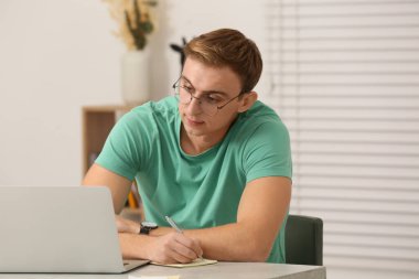 Young man with laptop writing in notebook at table indoors