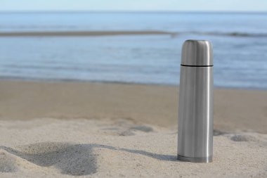 Metallic thermos with hot drink on sandy beach near sea, space for text