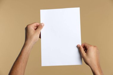 Man holding sheet of paper on beige background, closeup. Mockup for design
