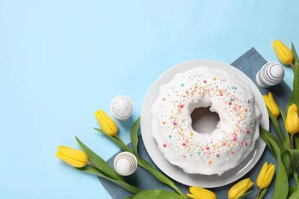 stock image Delicious Easter cake decorated with sprinkles near beautiful tulips and painted eggs on light blue background, flat lay. Space for text