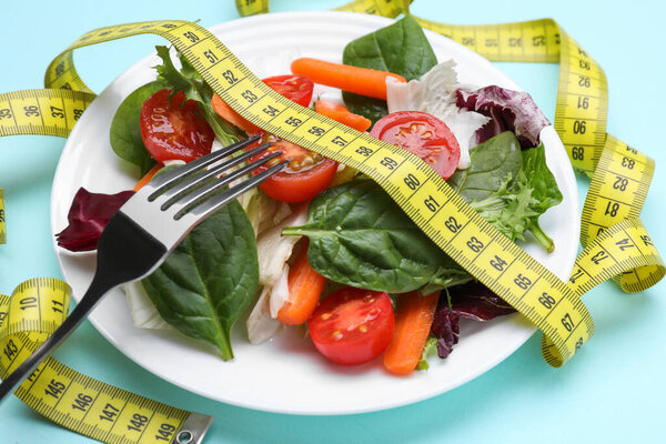 Measuring tape, vegetable salad and fork on light blue background, closeup. Weight loss concept