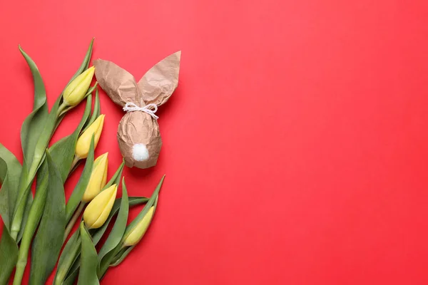 Easter bunny made of kraft paper and egg near beautiful tulips on red background, flat lay. Space for text