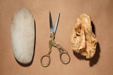 Rock, crumpled paper and scissors on light brown background, flat lay