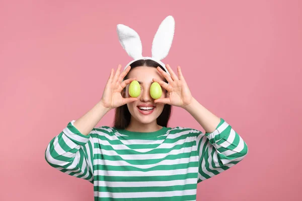 stock image Happy woman in bunny ears headband holding painted Easter eggs near her eyes on pink background