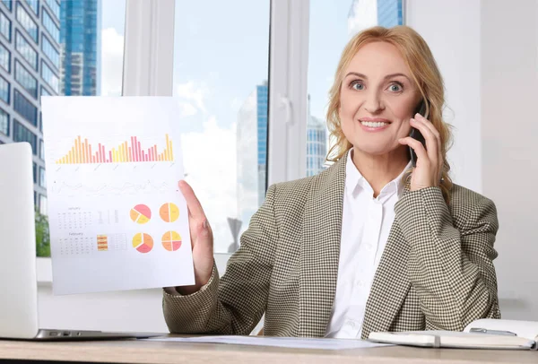 女性の上司はスマートフォンで話し オフィスのデスクで統計のある論文を見せています 成功した実業家 — ストック写真