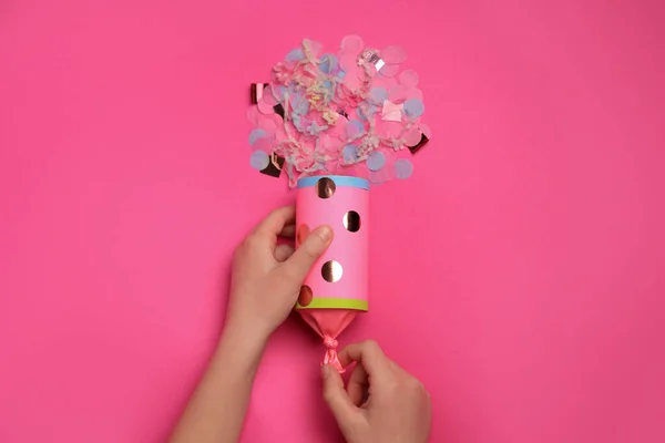 Woman holding party popper with serpentine and confetti on pink background, top view