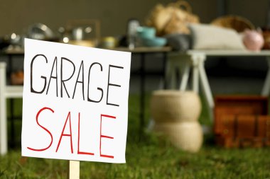 Sign Garage sale written on cardboard near tables with different stuff outdoors, closeup. Space for text