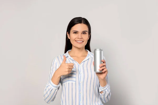 Stock image Beautiful young woman holding tin can with beverage and showing thumb up on light grey background