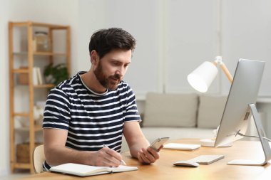 Ev işyerinde. Akıllı telefon kullanan ve odasındaki ahşap masada notlar alan bir adam.