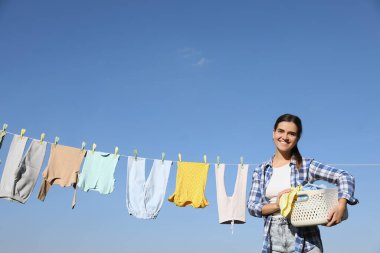 Mavi gökyüzüne karşı kurumak için çamaşır ipinin yanında bebek kıyafetleriyle gülümseyen kadın.