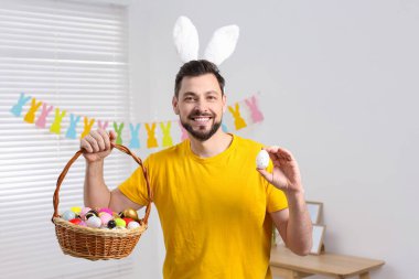 Tavşan kulaklı mutlu adam elinde hasır sepet, içinde boyanmış Paskalya yumurtaları tutuyor.