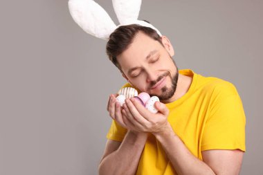 Happy man in bunny ears headband holding painted Easter eggs on grey background. Space for text