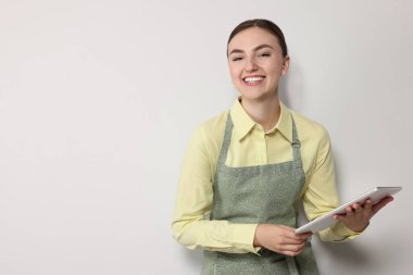 Beautiful young woman in clean apron with tablet on light grey background. Space for text