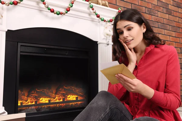 Emotional woman sitting near fireplace with greeting card indoors
