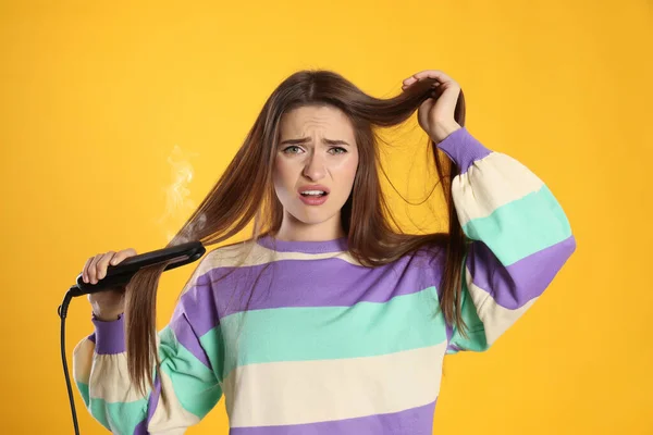 stock image Stressed young woman with flattening iron on yellow background. Hair damage