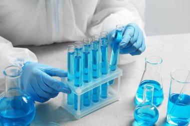 Bilim adamı laboratuardaki beyaz masada açık mavi sıvıyla test tüpü alıyor.