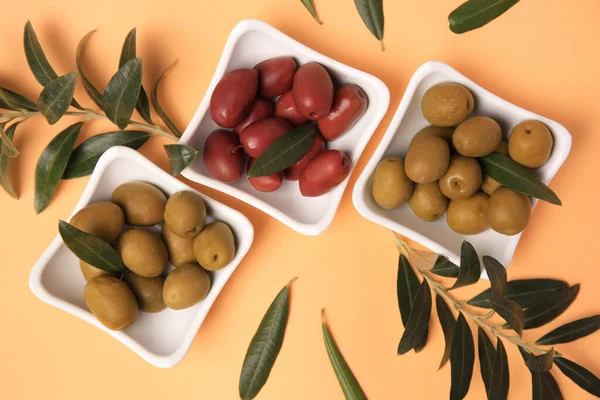 Different fresh olives and green leaves on pale orange background, flat lay