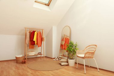 Stylish clothes rack, mirror and wicker chair in attic room. Interior design