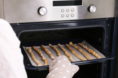 Woman taking baking sheet with homemade breadsticks out of oven, closeup. Cooking traditional grissini