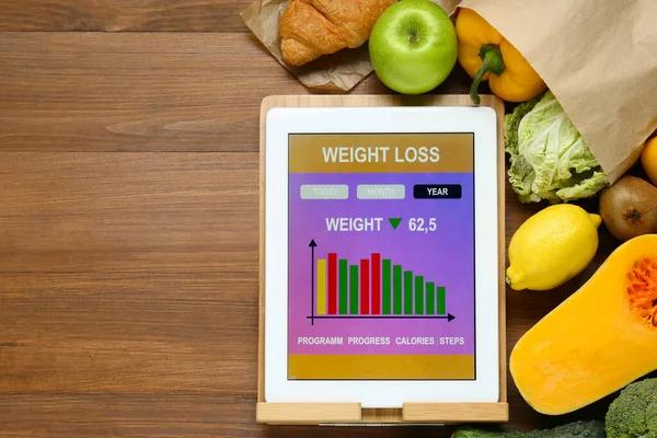 Tablet with weight loss calculator application and food products on wooden table, flat lay. Space for text