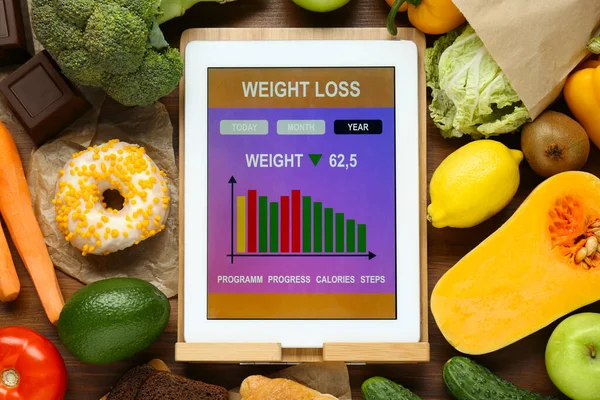 Tablet with weight loss calculator application and food products on wooden table, flat lay